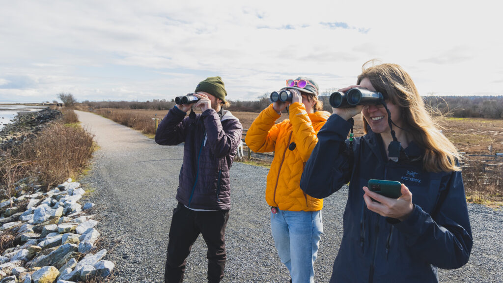 Three women are looking through binoculars and one is holding a cell phone.