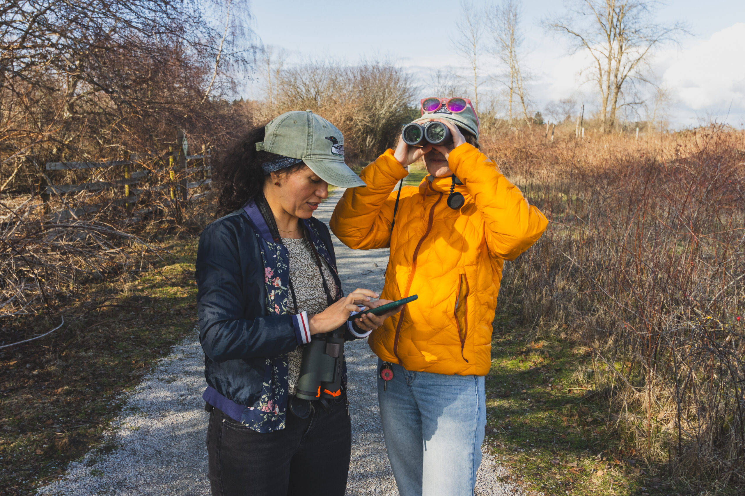 Two birdwatchers stand on a trail. One is looking into the distance through binoculars, and the other is tapping a mobile phone screen..