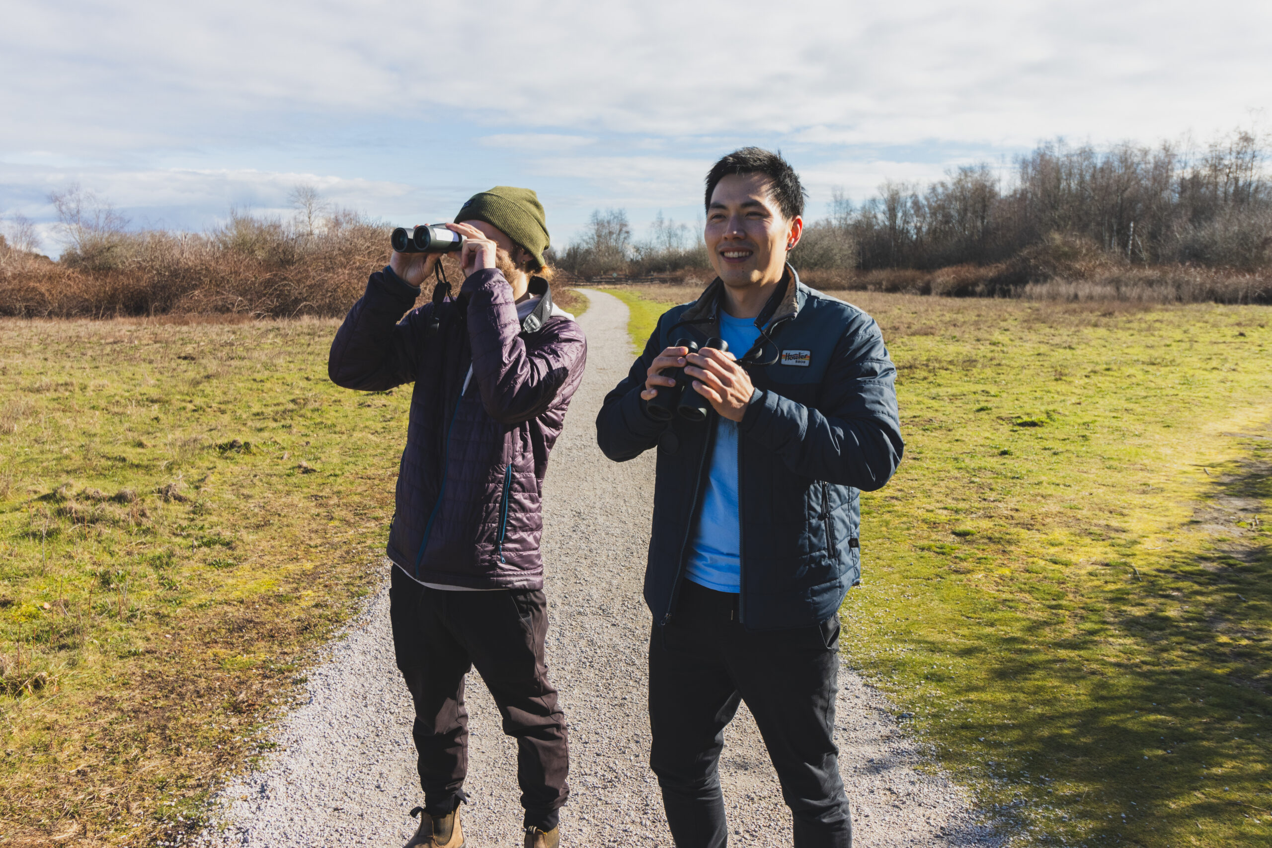 Two birders stand on a trail, looking into the distance through binoculars.
