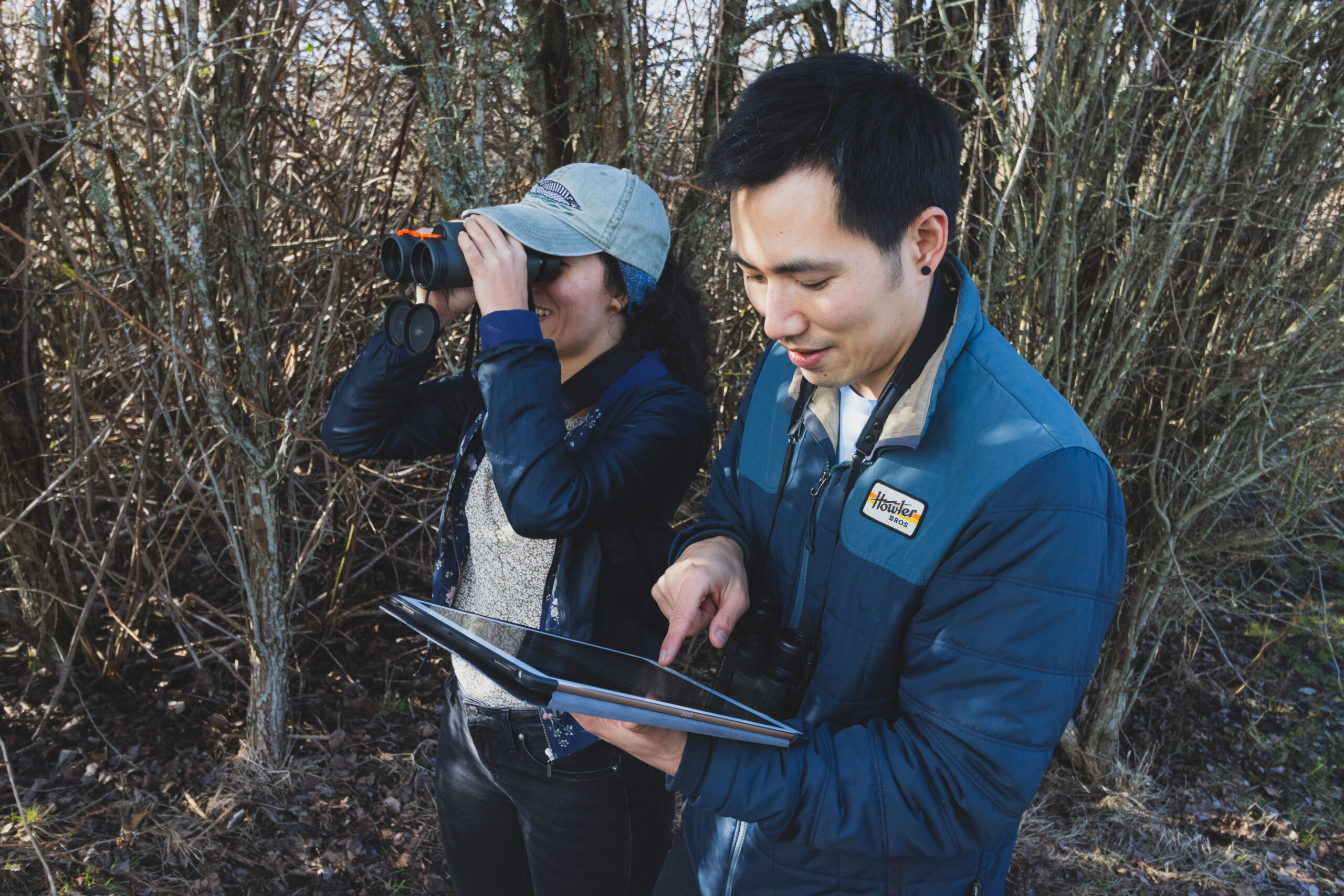 Two birdwatchers stand on a trail. One is looking into the distance through binoculars, and the other is tapping a tablet screen..